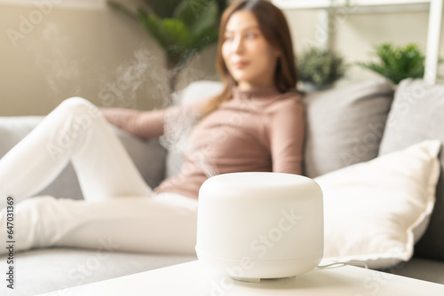 Modern air humidifier during relax or rest, happy blurred asian young woman, girl enjoying aromatherapy steam scent from essential oil diffuser comfortable in living conditions room,apartment at home