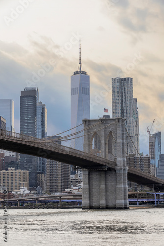 The iconic Brooklyn Bridge crosses the East River leading into Manhattan, with sun beams shining down on New York City's skyscrapers.