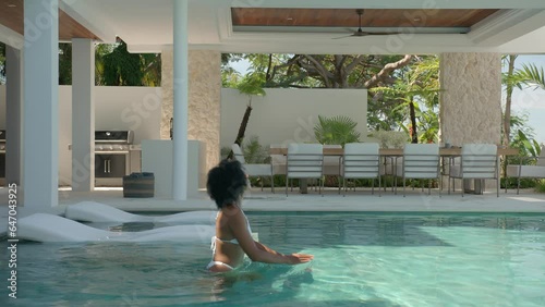 Beautiful black bikini woman walks into pool side view, girl enjoys her time in tropical lux hotel, sunny day at spa concept.