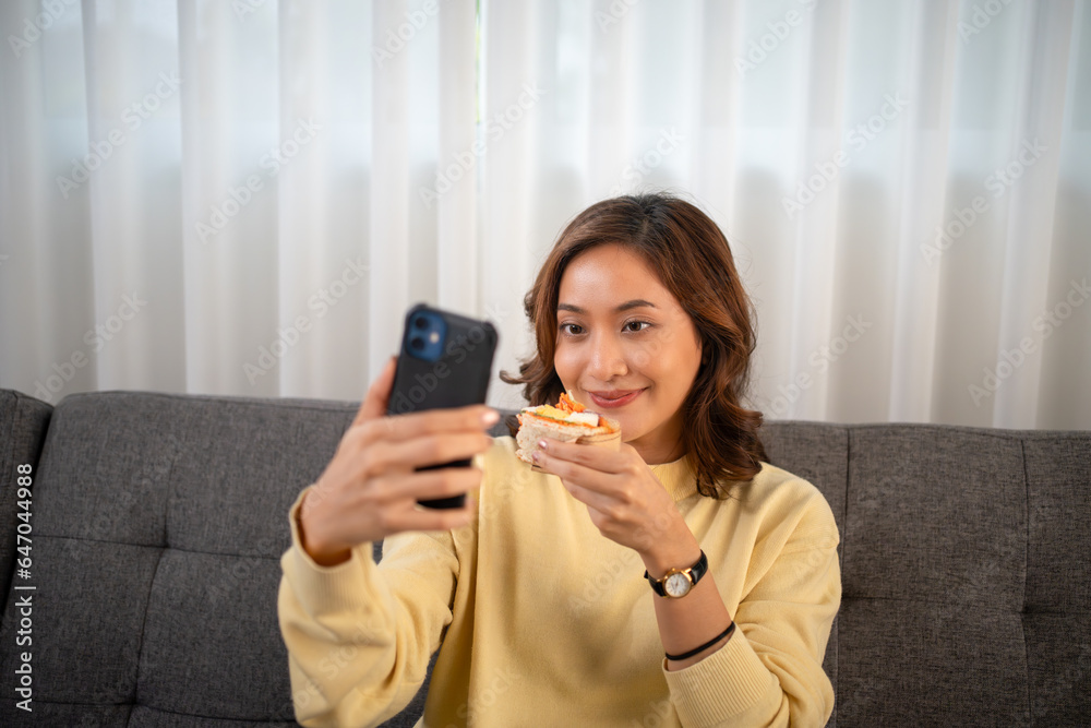Asian woman using phone video call with friend while eating sandwich at home Asian woman using phone video call with friend while eating sandwich at home