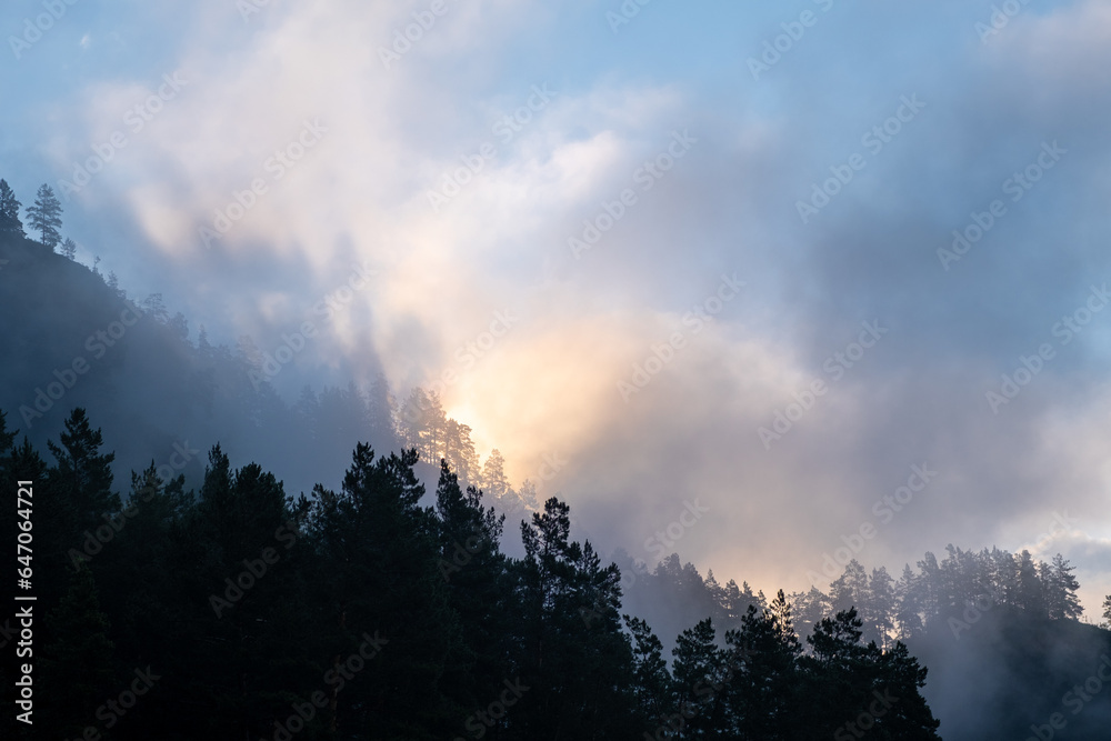 Mountains and peaks in fog at sunrise or sunset. Sun from behind the mountain. Amazing view of mountains and forest landscape with cloudy skies Altai mountains. Mountains and peaks in fog at sunrise