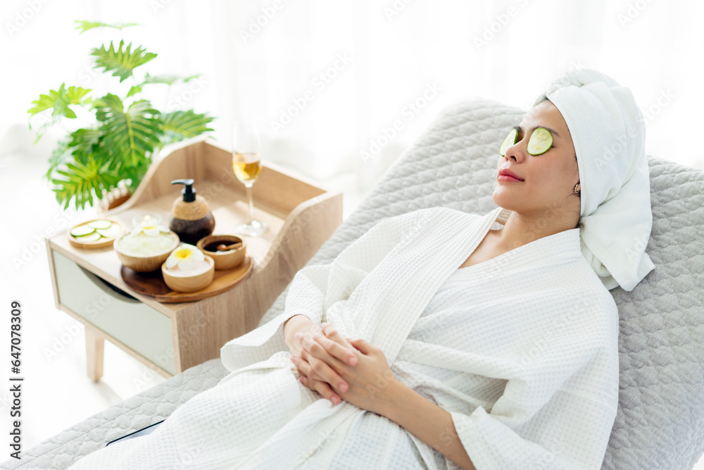 Relax young Asian woman in bathrobe, towel on a head, lying on the sofa bed with sliced cucumbers on her eyes and beauty treatment items for spa procedures such as essential oil and salt massage. - obrazy, fototapety, plakaty 