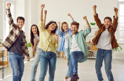 Funny happy overjoyed group of friends celebrating success holding hands up and looking at the camera at home. Group of smiling people students in casual clothes standing together enjoying party.