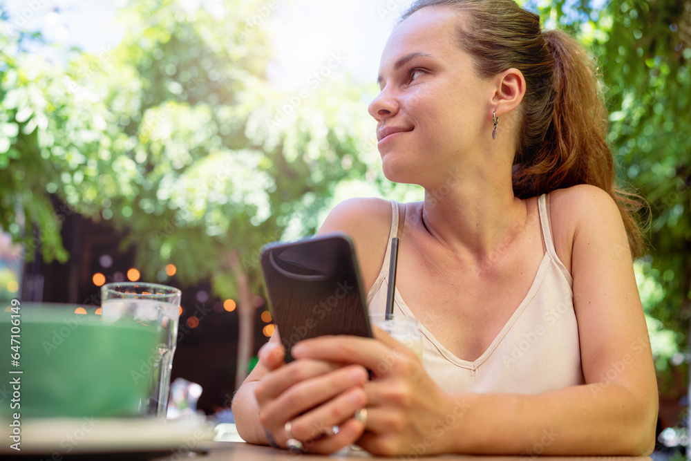 Low angle view young brunette woman sitting in open air cafe in summer with mobile phone in her hands. Low angle view young brunette woman sitting in open air cafe in summer with mobile phone in her hands.
