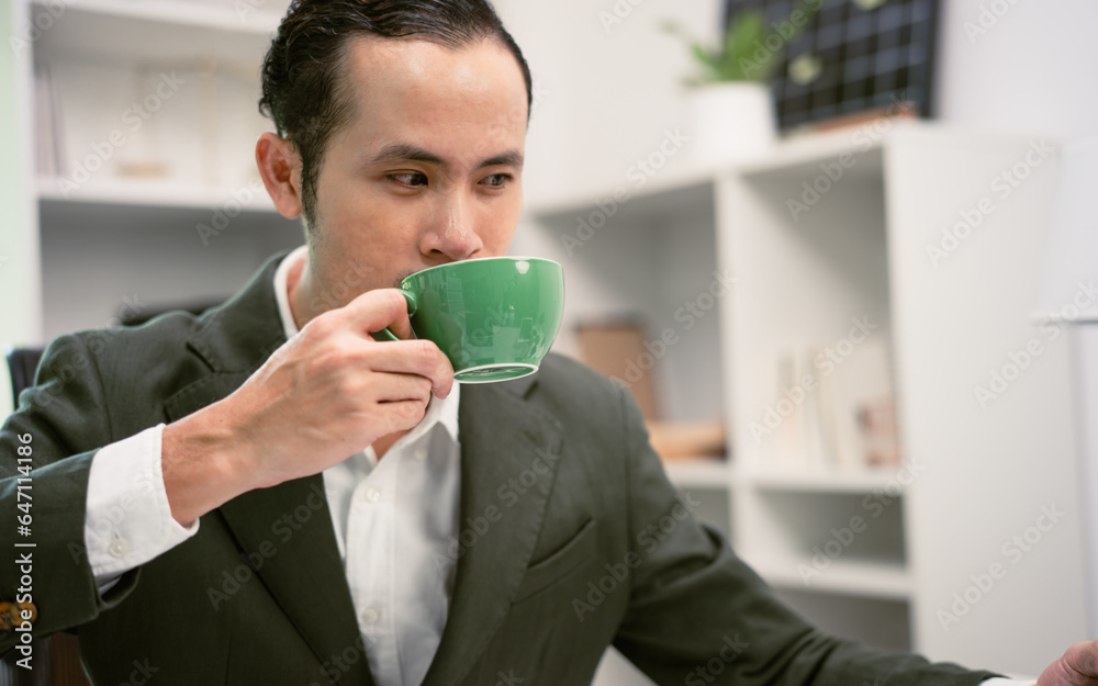 The manager of an Asian man sipping coffee on his desk in a radiant manner.
