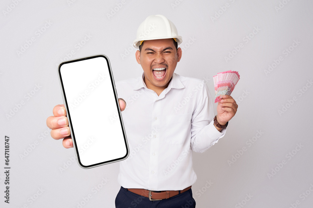 Smiling happy young Asian architect engineer employee man in a formal shirt helmet holding smartphone with blank screen and money isolated on white background. People work on architecture project Smiling happy young Asian architect engineer employee man in a formal shirt helmet holding smartphone with blank screen and money isolated on white background. People work on architecture project