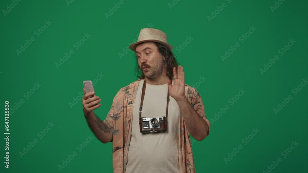 Man traveller in casual clothing and straw hat talking with friend by a video call on smartphone, showing new retro film camera he bought. Isolated on green background. Man traveller in casual clothing and straw hat talking with friend by a video call on smartphone, showing new retro film camera he bought. Isolated on green background.