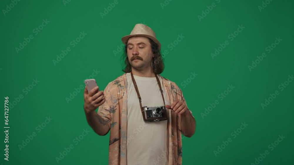 Man traveller in casual clothing and straw hat with old film camera taking selfie on smartphone, smiling at the camera. Isolated on green background. Man traveller in casual clothing and straw hat with old film camera taking selfie on smartphone, smiling at the camera. Isolated on green background.