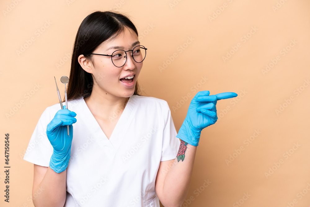 Dentist Chinese woman holding tools isolated on beige background pointing finger to the side and presenting a product Dentist Chinese woman holding tools isolated on beige background pointing finger to the side and presenting a product