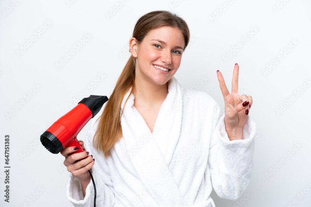 Young caucasian woman holding hairdryer isolated on white background smiling and showing victory sign Young caucasian woman holding hairdryer isolated on white background smiling and showing victory sign