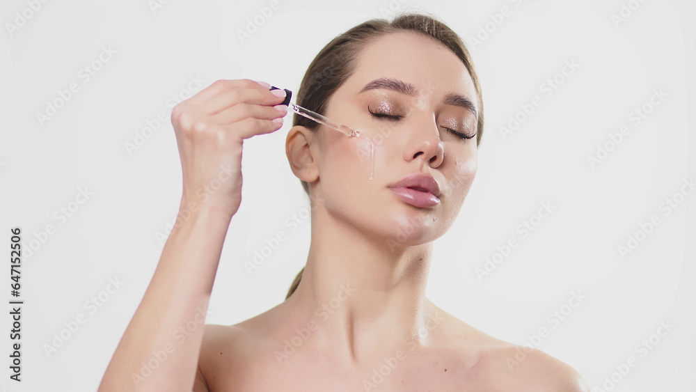 Close-up studio portrait of a young woman, isolated on a light background, applying serum from a pipette. Natural organic cosmetic product for facial skin care and skin regeneration.
