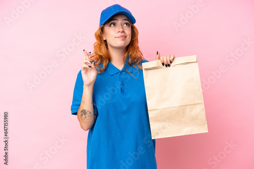 Young caucasian woman taking a bag of takeaway food isolated on pink background with fingers crossing and wishing the best