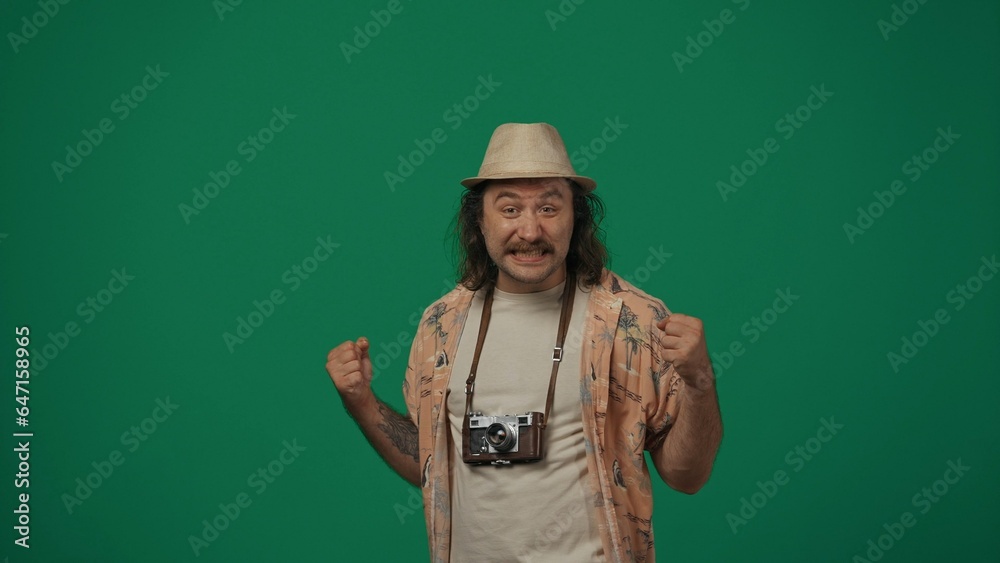 Man traveller in casual clothing and straw hat with old film camera pumping his fists in the air, happy face expression. Isolated on green background. Man traveller in casual clothing and straw hat with old film camera pumping his fists in the air, happy face expression. Isolated on green background.