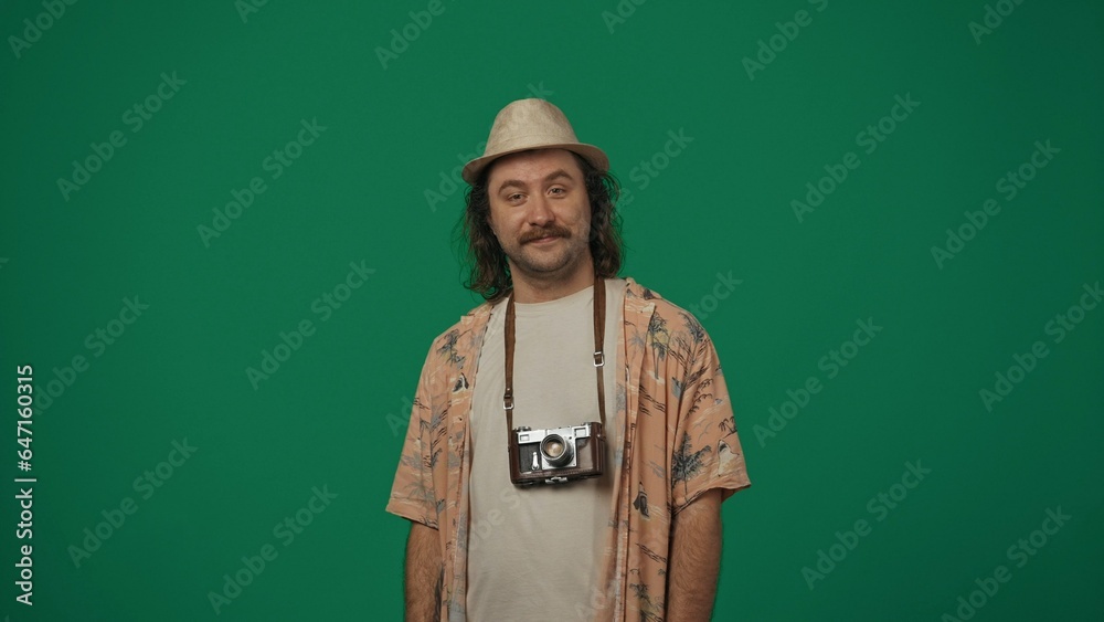 Man traveller in casual clothing and straw hat with old film camera over neck posing, looking directly at the camera, smiling. Isolated on green background.