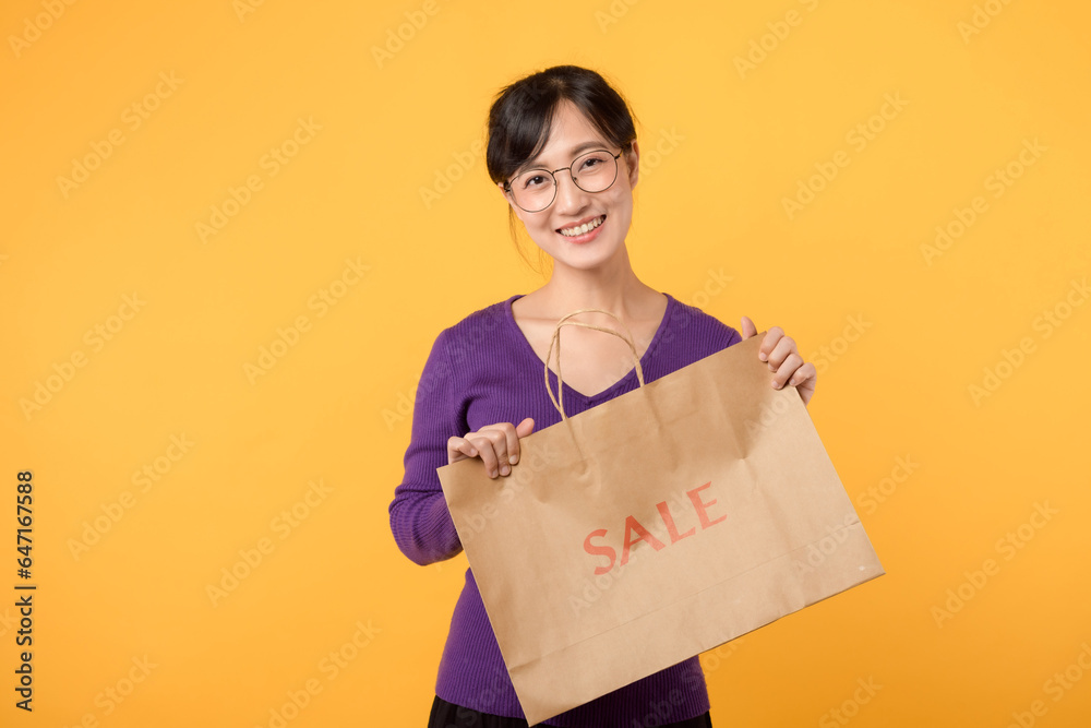 Asian young beautiful woman 30s wearing purple shirt holding paper shopping bag, presenting environmental sustainable packages, smiling with happiness isolated on yellow background with copy space. Asian young beautiful woman 30s wearing purple shirt holding paper shopping bag, presenting environmental sustainable packages, smiling with happiness isolated on yellow background with copy space.