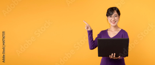 Asian female teacher 30s cute smile holding laptop pointing finger to free copy space, Wearing purple sweater shirt and standing in a yellow studio setting. Exchange education advertising concept.