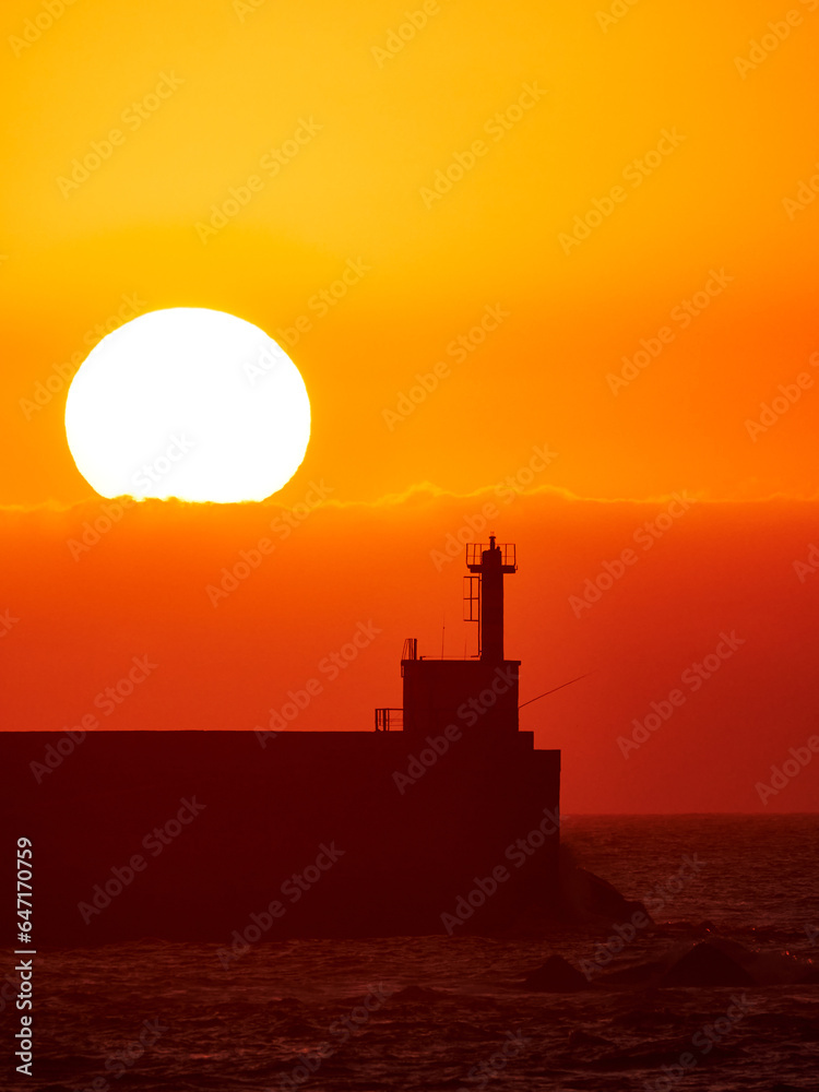 Distant beacon on pier rising above sea water against big bright sun setting at horizon