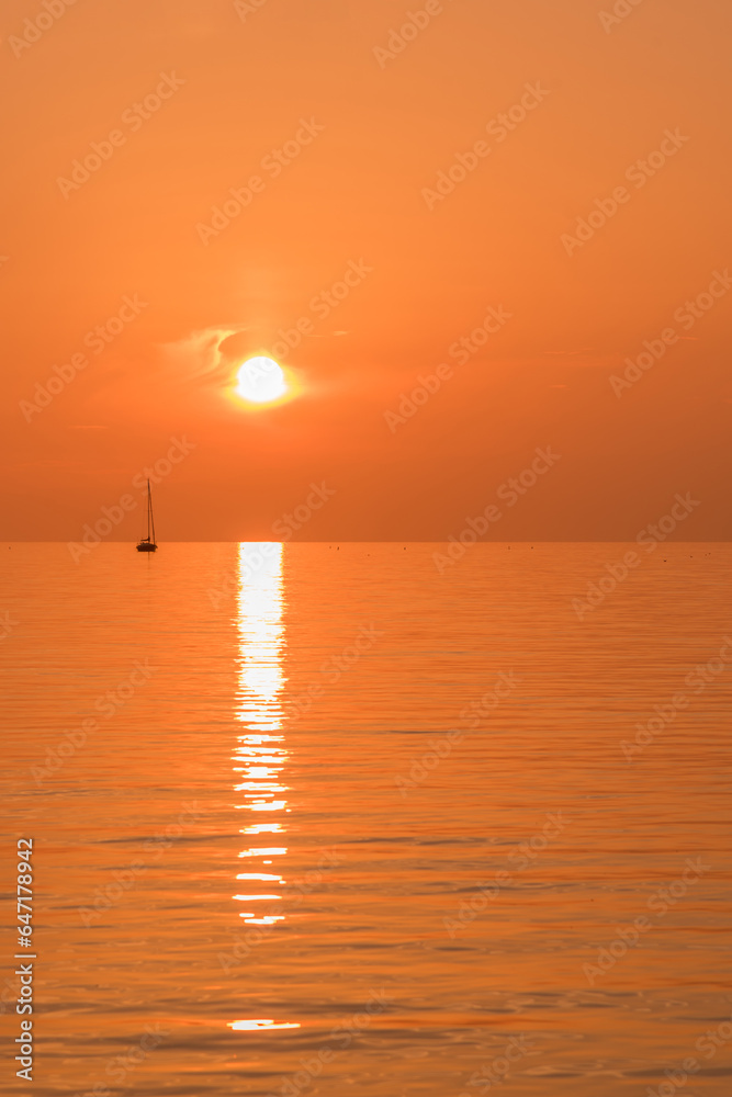 View of a sailboat sailing on a lake during sunset with an orange sky. View of a sailboat sailing on a lake during sunset with an orange sky.