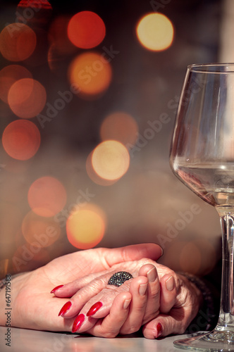Young daughter holding old mother's hand. Close up of mom and daughter hands next to a wine glass and bokeh lights as background. Date with mom.