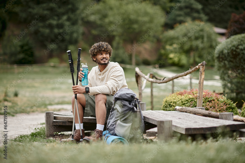 Fototapeta premium cheerful indian hiker holding trekking poles and sports bottle near backpack in forest