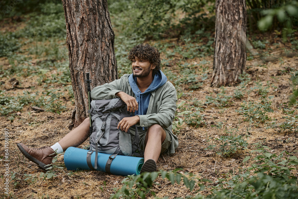 carefree young indian tourist resting while sitting near backpack on ground in forest