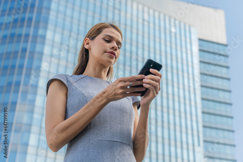 Business on the move. Young caucasian white businesswoman using smartphone while commuting in a central business district against a background of modern skyscraper cityscape.