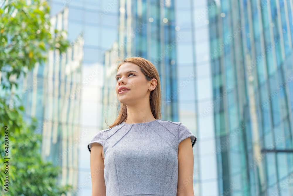Fototapeta premium Portrait of a young and successful white caucasian business woman standing in front of a modern cityscape of office buildings.