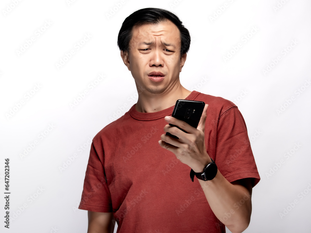 An Asian Indonesian man wearing a maroon-colored shirt appears to be sleepy while looking at his smartphone, isolated against a white background. An Asian Indonesian man wearing a maroon-colored shirt appears to be sleepy while looking at his smartphone, isolated against a white background.