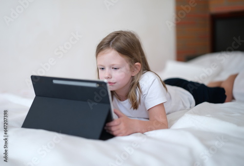 Little girl carefully looks at tablet while lying on bed. Mobile learning apps for kids concept