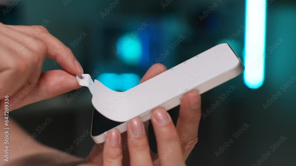 Close-up side view of unrecognizable man removing protective film from new smartphone on bokeh light background in dark room. Closeup hands with accuracy taking off transparent plastic protective film