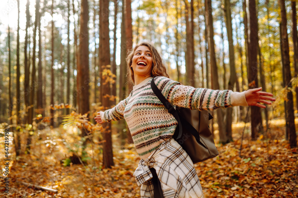Tourist with a hiking backpack, hat walks along a path in the autumn forest. Beautiful woman enjoys a sunny day in nature, feels freedom and breathes fresh air, explores nature. - obrazy, fototapety, plakaty 