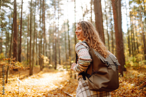 Tourist with a hiking backpack, hat walks along a path in the autumn forest. Beautiful woman enjoys a sunny day in nature, feels freedom and breathes fresh air, explores nature.