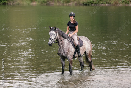 Gray horse with a female rider in the saddle walking through river water. Ecotourism and equestrian recreation sport concept. © 24K-Production