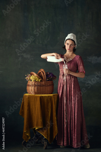 Elegant young woman in renaissance dress, maid pouring milk into glass against dark vintage background. Peasant girl. Concept of history, comparison of eras, beauty, art, creativity photo