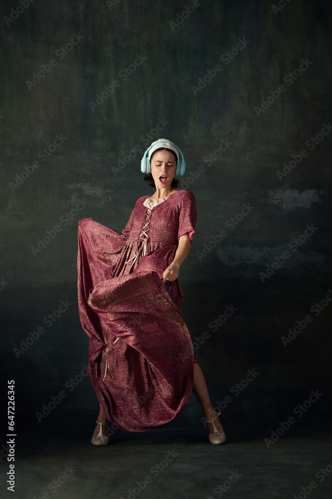 Medieval woman in elegant renaissance dress, maid listening to music in headphone and dancing against vintage green background. Concept of history, comparison of eras, beauty, art, creativity