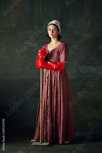 Beautiful, elegant woman, medieval maid in dress standing with mop in big red rubber gloves against dark vintage background. Concept of history, comparison of eras, beauty, art, creativity