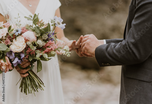 Close up of couple's hands exchanging rings at an outdoor ceremony on a pebble beach with flowers