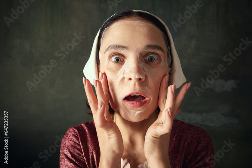 Obraz na plátne Close-up of young woman in image of medieval person, maid, taking care after skin with face mask against dark vintage background