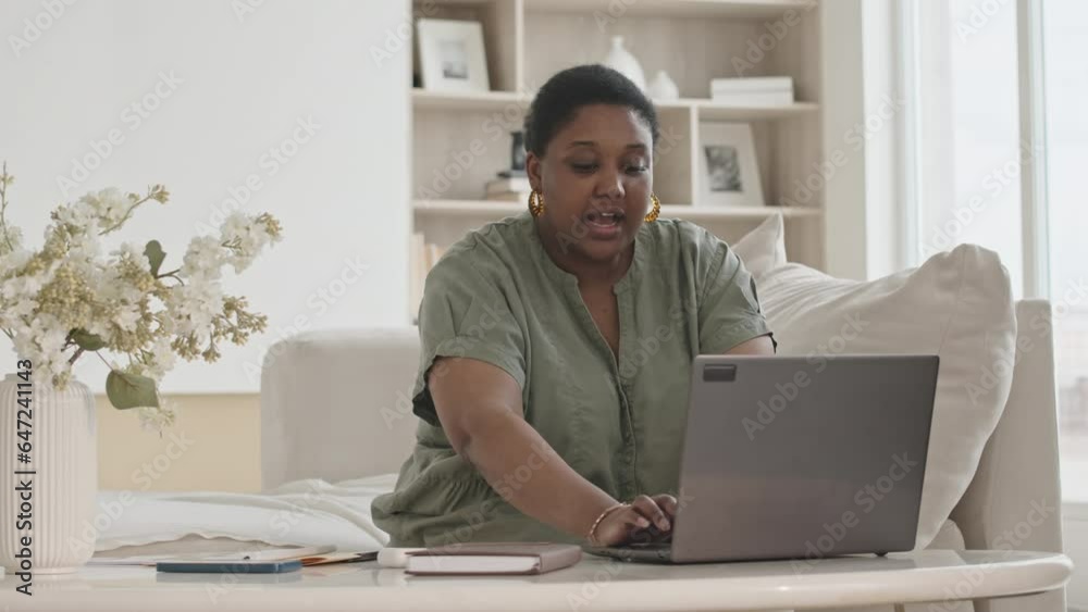 Medium shot of African American woman sitting in living room, typing on laptop and looking through documents while working from home Medium shot of African American woman sitting in living room, typing on laptop and looking through documents while working from home