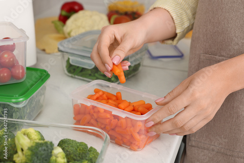 Woman putting carrots into container with fresh vegetables at white marble table in kitchen, closeup. Food storage