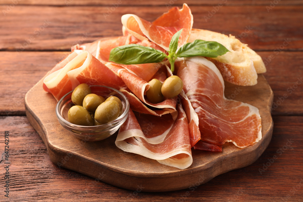 Slices of tasty cured ham, olives, bread and basil on wooden table, closeup