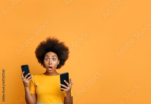 Young surprised and confused black woman holding two cellphones in front of a yellow background 