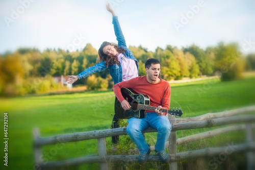 Romantic happy cute caucasian couple playing guitar and singing songs sitting on wooden fence in meadow in outdoor nature park