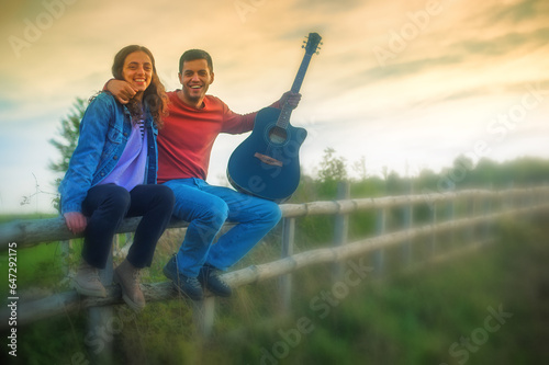 Romantic happy cute caucasian couple playing guitar and singing songs sitting on wooden fence in meadow in outdoor nature park