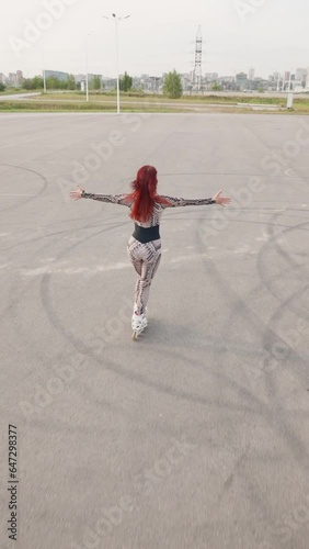 Vertical Video, Slim Woman with Red Hair Has Fun Rollerblading on an Asphalt Parking Lot. Happy Girl Roller-Skates and Raises Her Arms to the Sides, Feeling Freedom and Wellbeing. Slow Motion.