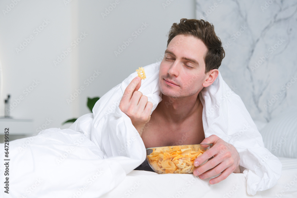 © Евгений Шемякин - Young man eating chips in bed, bedroom. Fast junk food.
