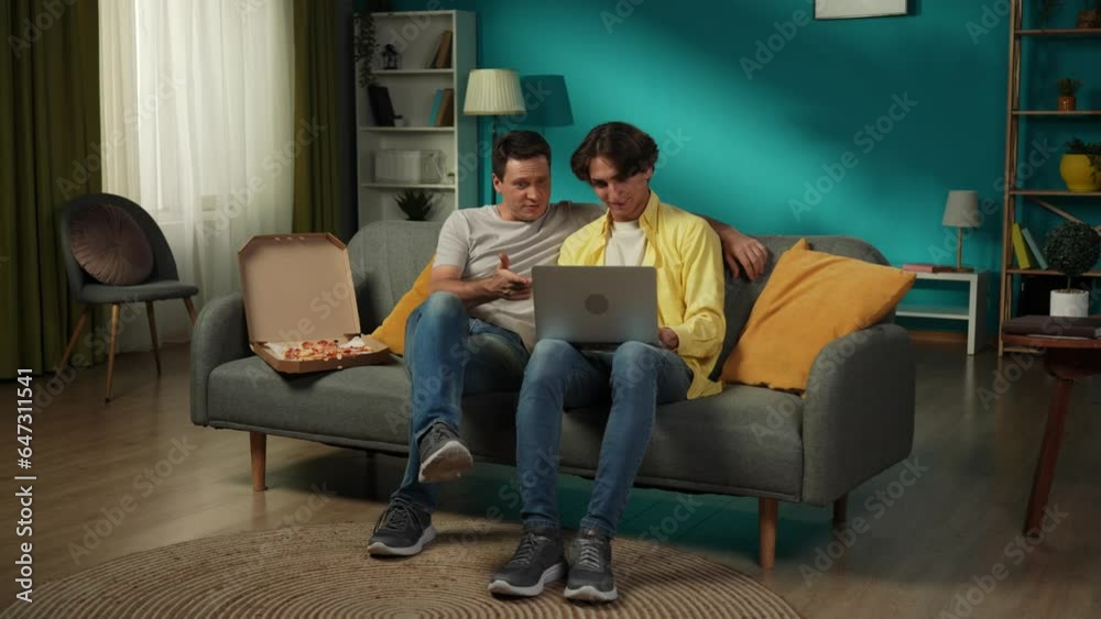 Full-size video of a homosexual couple at home. They are sitting on the couch, watching photos on a laptop, eating pizza, hugging and talking warmly to each other. Full-size video of a homosexual couple at home. They are sitting on the couch, watching photos on a laptop, eating pizza, hugging and talking warmly to each other.