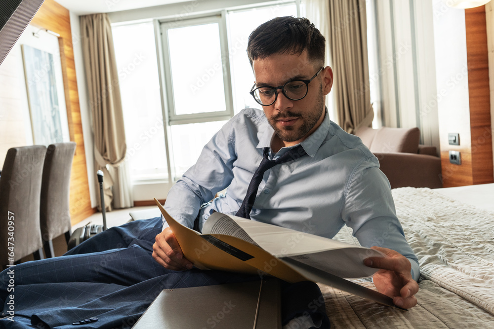 Fototapeta premium The businessman enters his hotel room, takes a seat, and then, with a sense of relief, sinks onto the hotel bed. The comfort of the room offers a well-deserved moment after a long day of meetings.