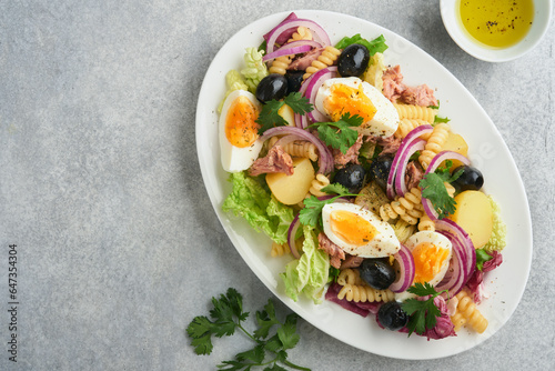 Tuna salad with pasta, eggs, potatoes, olives, red onions and sauce in white plate on old light gray concrete table background. Nicoise salad. French cuisine. Top view. Flat lay.