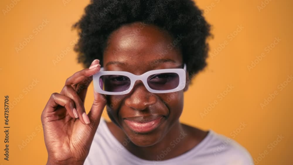 An African American young woman with an Afro hairstyle and glasses stands in a sweater on a bright orange background looking at who is there while taking off her glasses. An African American young woman with an Afro hairstyle and glasses stands in a sweater on a bright orange background looking at who is there while taking off her glasses.